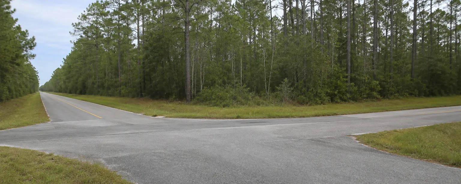 A quiet paved crossroads where two rural two-lane roads meet, surrounded by pine woods on every corner